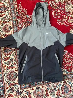 Nike tvåfärgad vindjacka med huva - Snygg vindjacka från Nike i svart och grått med dragkedja framtill och två fickor med dragkedja. Jackan har en justerbar huva och sportig design med Nike-logga på bröstet. Knappt använd med infa synliga skadliga defekter! Köptes hösten 2025 har digital kvitto nyköpt för 1000kr