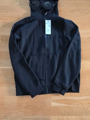 Svart hoodie från C.P. Company - Svart hoodie från C.P. Company med dragkedja och ikoniska glasögon i huvan. Broderad logga på bröstet och stora fickor framtill. Den är i helt nytt skick och aldrig använd. Den har alla etiketter kvar och har skyddsplasten kvar på glasögonen. Den är i storleken L men är som en M i storleken. 