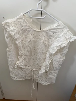 Vit broderad blus från Gina Tricot - Söt vit blus från Gina Tricot med broderade detaljer och volangkant längs ärmhålen. Blusen har knytband i midjan och är tillverkad i ett lätt bomullstyg med hålmönster. Perfekt för en somrig och romantisk look.