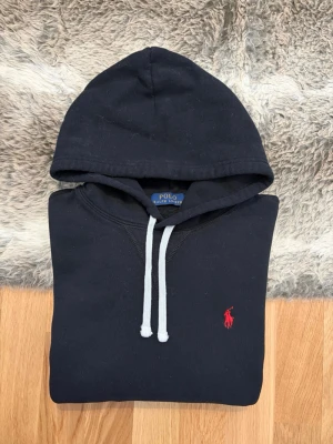 Svart Polo Ralph Lauren Lauren Hoodie - Storlek M - Hej! 👋 Säljer en riktigt snygg och klassisk svart Polo Ralph Lauren fleece-hoodie i storlek M. Den har den ikoniska röda pony-loggan broderad på bröstet – en tidlös färgkombination som alltid är rätt! 🐎 Tröjan är i mycket gott skick och välvårdad. 100% äkta med både tvättlappar och QR-kod kvar för enkel verifiering ✅ Ett högkvalitativt basplagg som sitter perfekt och håller stilen år efter år. Fraktar snabbt! 🚛📦💬💯 