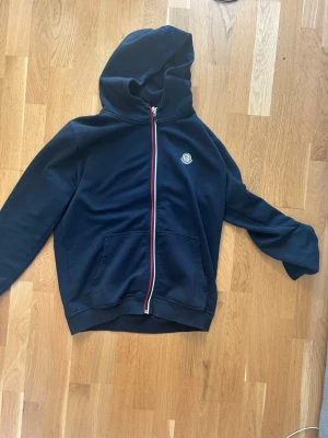 Mörkblå Moncler hoodie med dragkedja - Snygg mörkblå hoodie från Moncler med hel dragkedja framtill och klassisk logga på bröstet. Hoodien har känguruficka och rödvit rand längs dragkedjan. Perfekt för en avslappnad och stilren look.