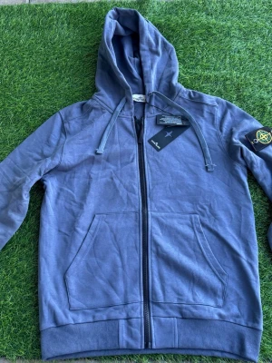 Blå hoodie från Stone Island - Snygg blå hoodie från Stone Island med dragkedja framtill och klassisk logotyp-patch på vänster ärm. Tröjan har huva med dragsko och två stora fickor framtill. Perfekt för en avslappnad streetwear-look.