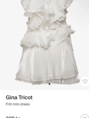 Vit volangklänning från Gina Tricot - Kort vit klänning från Gina Tricot med volangdetaljer framtill och omlottkänsla. Tillverkad i ett glansigt material som ger en lyxig look. Perfekt för dig som vill sticka ut med en feminin och trendig stil.