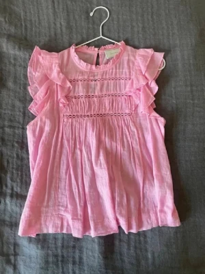 Rosa volangblus från Neo Noir - Söt rosa blus från Neo Noir med volangdetaljer längs ärmhålen och smockad överdel. Blusen är ärmlös och har rund halsringning samt knapp i nacken. Tillverkad i ett lätt, luftigt tyg som ger en romantisk känsla.