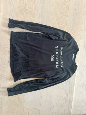 Svart långärmad topp Acne Studios - Svart långärmad topp från Acne Studios med tryck i vitt framtill där det står 'Acne Studios STOCKHOLM 1996'. Tunn och mjuk kvalitet, rund halsringning och rak passform. Perfekt för en stilren och modern look.