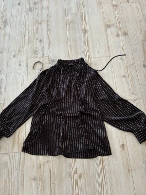 Svart blus med ränder och knytband - Snygg svart blus med tunna vertikala ränder i vitt och rosa. Blusen har lång ärm med resår vid ärmslut och ett dekorativt knytband vid halsen. Perfekt för en trendig och avslappnad look.från Stockholm MQ