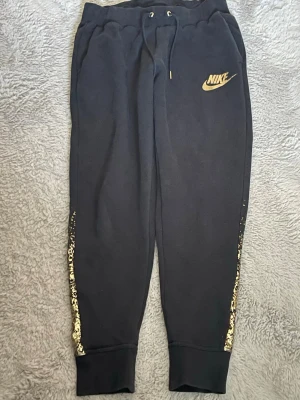 Svarta Nike mjukisbyxor med guldtryck - Svarta mjukisbyxor från Nike med guldfärgat logotryck på låret och coola guldfärgade detaljer längs benen. Byxorna har resår och dragsko i midjan samt muddar vid bensluten. Perfekta för en avslappnad och trendig look.
