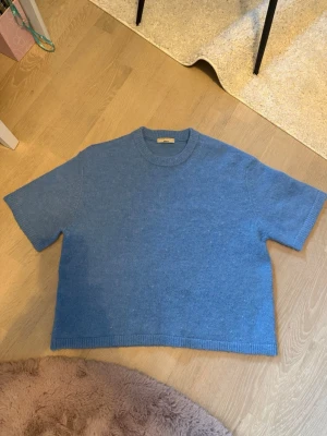 Blå stickad t-shirt från Gina Tricot - Kortärmad stickad t-shirt i en mjuk blå nyans från Gina Tricot. Tröjan har rund halsringning och ribbade kanter vid ärmslut och nederkant. Perfekt för lager-på-lager och chill stil. Materialet ser ut att vara ull eller ullblandning. Är egentligen i mycket bra skick men är dock lite nopprig men det syns inte så mycket. 