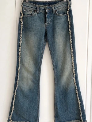 Blå bootcut jeans med fransade kanter - Säljer ett par blå bootcut jeans med råa, fransade kanter längs sidorna och nederkanten. Jeansen har klassisk femficksdesign, normal midja och är tillverkade i denim. Perfekta för dig som gillar en unik och trendig look.