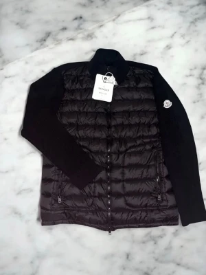 Svart dunjacka från Moncler - Snygg svart dunjacka från Moncler med quiltad front, två dragkedjeförsedda fickor och Moncler-logga på ärmen. Jackan har hög krage och raka ärmar i mjukare material, perfekt för kalla vinterdagar. Stängs med dragkedja framtill.