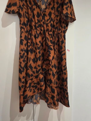 Leopardmönstrad omlottklänning Shein - Snygg leopardmönstrad omlottklänning från Shein Curve i bruna och svarta toner. Klänningen har korta ärmar, volangdetalj framtill och markerad midja. Perfekt för dig som vill sticka ut med ett djurmönster och en trendig look.