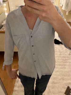 Ljusblå randig henley-skjorta - Ljusblå henley-skjorta med diskreta ränder och knappar framtill. Skjortan har en bröstficka och trekvartsärmar med knyt vid ärmsluten. Perfekt för en avslappnad och stilren look.