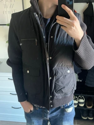 Corneliani dunväst med stickade ärmar - Italienskt lyx märke ”Corneliani ID”. Nypris ligger på 800€ - 1300€. Inga syn på användning. Snygg svart dunväst med stickade ärmar och hög krage. Västen har två stora fickor med lock och knappar, samt en bröstficka med dragkedja. Skickar till sverige🇸🇪
