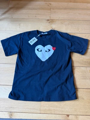 Svart Comme des Garçons Play t-shirt - Svart t-shirt från Comme des Garçons Play med stort vitt hjärta och ögon på bröstet samt ett litet rött hjärta. Klassisk rund hals och korta ärmar. Perfekt statement-plagg för dig som gillar streetwear och ikoniska prints. Glöm inte att den kan passa en storlek mindre asså M. Pris kan diskuteras.