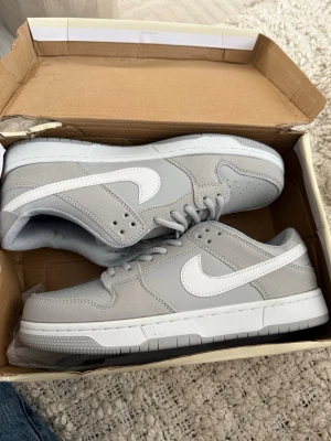 Nike SB Dunk Low grå/vit sneakers - Nike SB Dunk Low sneakers i stilren grå och vit färg. Skorna har klassisk låg siluett, vit swoosh och slitstark ovandel i mocka och textil. Perfekta för dig som gillar streetwear och skate. Levereras i originalkartong.