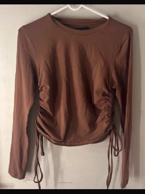 Brun långärmad topp  - Snygg brun långärmad topp från Pieces med trendig snörning i sidorna som ger en cool, rynkad effekt. Toppen har rund halsringning och är tillverkad i mjuk bomull, perfekt att styla med jeans eller kjol.