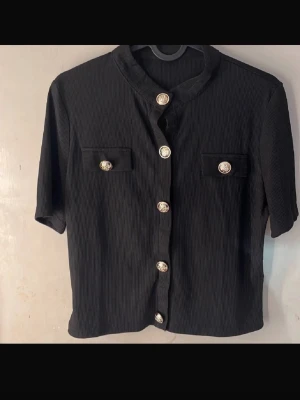 Svart topp med guldknappar - Stilren svart topp med struktur och dekorativa guldknappar framtill samt på bröstfickorna. Skjortan har korta ärmar och rak passform, perfekt för en trendig look.