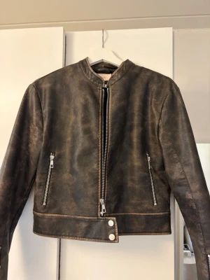 Vintage skinnjacka - Snygg brun skinnjacka med cool asymmetrisk dragkedja och två fickor med silverfärgade dragkedjor. Jackan har en kortare passform och rund halsringning samt dekorativt bälte med knappar nedtill.