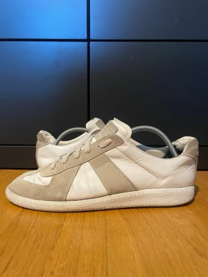 Vita och grå sneakers från Maison Margiela - Snygga sneakers från Maison Margiela i vitt och grå läder och mocka. Klassisk låg modell med vita snören och gummisula. Ikonisk design med paneler i mocka och diskret logga på plösen. Perfekta för en clean och stilren look.