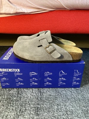 Grå mockatofflor Birkenstock Boston - Säljer ett par gråa Birkenstock Boston tofflor i mocka med klassisk design och justerbart spänne i metall. De har en bekväm korksula och är perfekta för en avslappnad stil. Modellen är smal läst och färgen är mörkgrå.