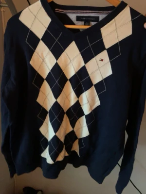 Mörkblå argyletröja Tommy Hilfiger - Snygg mörkblå stickad tröja från Tommy Hilfiger med klassiskt argylemönster i vitt och marinblått framtill. V-ringad modell med långa ärmar och diskret logga på bröstet. Perfekt för en preppy stil.