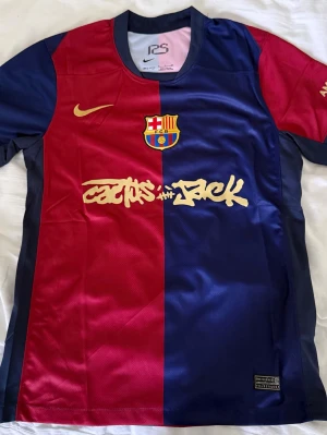 Barça x Cactus Jack matchtröja Nike - Säljer en FC Barcelona matchtröja i samarbete med Cactus Jack och Nike. Tröjan är röd och blå med klubbens emblem på bröstet, Nike-logga och Cactus Jack-tryck i guld. Materialet är lätt och andas, perfekt för sport och träning. UNHCR-logga på ryggen.