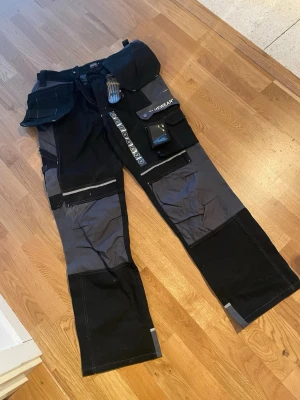 Svarta och grå arbetsbyxor Bluewear - Tuffa arbetsbyxor från Bluewear i svart och grått med flera praktiska fickor, hammarhållare och förstärkta knän. Perfekta för dig som vill ha funktionella och slitstarka byxor med smarta detaljer och reflexpartier. Materialet är tåligt och anpassat för arbete. 