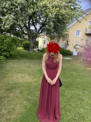 Vinröd balklänning med öppen rygg - Säljer en superfin vinröd långklänning med superfin passform. Den går in i midjan vilket gör sen så smickrande. Klänningen har halterneck med dekorativa stenar och en snygg öppen rygg med tunna band. Perfekt för bal eller student. Materialet känns lätt och faller fint. Den har några fläckar nertill som syns på första bilden men jag har inte försökt få bort dem.