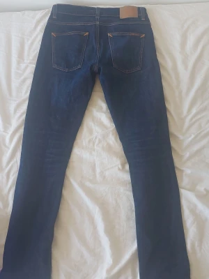 Mörkblå jeans från Nudie Jeans - Snygga mörkblå jeans från Nudie Jeans med klassisk femficksdesign och kontrastsömmar i orange. Jeansen har rak passform och är tillverkade i denim av bomull. Perfekta för en stilren och avslappnad look.