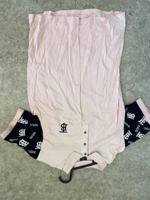 Ljusrosa henley med svarta ärmar - Snygg ljusrosa henley från LBDP med svarta korta ärmar som har vitt logomönster. Skjortan har knappar framtill och ribbad krage. Perfekt för dig som vill sticka ut med en unik stil.