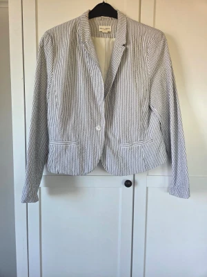 Randig kavaj från Lindex, vit och marinblå  - Snygg kavaj från Lindex med blå och vita ränder. Klassisk modell med en knapp framtill, två fickor och helfodrad insida. Perfekt för en stilren look. Materialet är lätt och passar bra till vår och sommar.