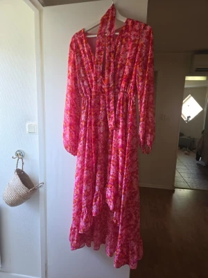 Blommig långklänning i rosa och orange - Långklänning med blommigt mönster i rosa och orange toner. Klänningen har långa ärmar, v-ringning och volangdetaljer nertill. Tillverkad i ett lätt och luftigt material, perfekt för varmare dagar. Snygg scarfdetalj eller skärp.