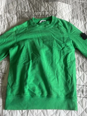 Grön sweatshirt från Stone Island - Snygg grön sweatshirt från Stone Island med klassisk svart logotyp-patch på vänster ärm. Tröjan har rund halsringning och är tillverkad i mjukt bomullsmaterial. Perfekt för en avslappnad och trendig stil. Köpt på NK Göteborg för 2800