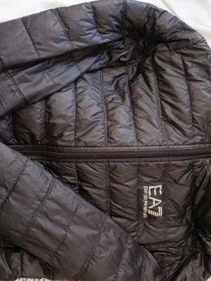 Svart pufferjacka från EA7 Emporio Armani - Snygg svart pufferjacka från EA7 Emporio Armani med huva och dragkedja framtill. Jackan har ett stilrent quiltat mönster och logga på bröstet. Perfekt för kalla sommar dagar och tillverkad i lätt, glansigt material som håller värmen.
