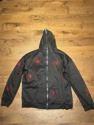 Svart hoodie med röda rhinestonespindlar - Svart hoodie med hel dragkedja och huva,  med röda spindelmotiv i glittrande stenar på bröstet, ärmarna och huvan. 