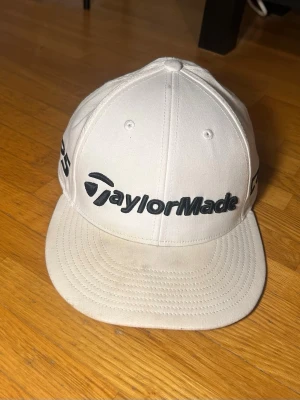 Vit TaylorMade keps med brodyr - Snygg vit keps från TaylorMade med svart broderad logga framtill och TP5-detaljer på sidorna. Klassisk böjd skärm och ventilerande hål upptill. Perfekt för golfbanan eller som sportig accessoar. Tillverkad i bomullstyg. Tour authentic.