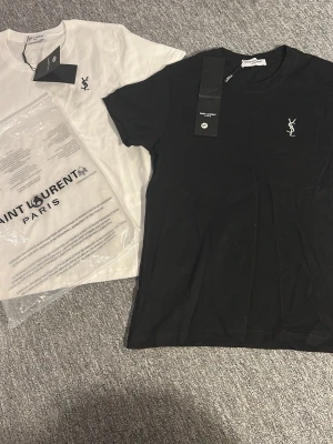 Saint Laurent svart och vit t-shirt - Två stilrena t-shirts från Saint Laurent, en svart och en vit, båda med klassisk YSL-logga broderad på bröstet. T-shirtsen har rund hals och korta ärmar, perfekta för en clean och trendig look. 2 för 899 annars 1 för 499. Skriv vid minsta fundering