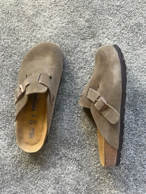 Birkenstock Boston taupe mockatofflor - Säljer ett par Birkenstock Boston tofflor i taupefärgad mocka med justerbart spänne i metall. Klassisk korksula och mjuk fotbädd för extra komfort. Perfekta för chill dagar hemma eller när du vill ha en avslappnad stil. Använda 3-5 gånger, låda medföljer ej