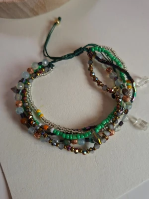 Färgglatt pärlarmband med flera rader - Snyggt armband med flera rader av små pärlor i grönt, blått, guld, silver och koppar. Armbandet har justerbar grön tråd och mixade former på pärlorna, vilket ger en unik och trendig look. Perfekt accessoar för att lyfta din outfit.