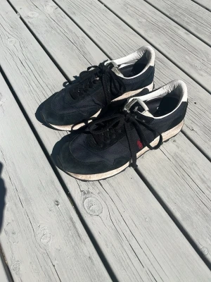 Svarta ralph louren sneakers med vit sula - Snygga svarta ralph louren sneakers med vit sula och klassisk låg profil. Ovandelen är i mesh och mocka, med svarta skosnören och diskret röd ralph louren detalj på sidan. Yttersulan har ett grovt mönster för bra grepp. Perfekta för en sportig och avslappnad stil.
