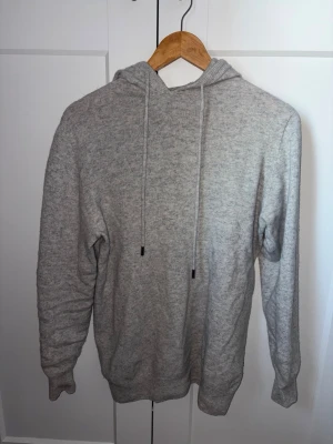 Ljusgrå stickad hoodie - Mysig ljusgrå stickad hoodie med huva och dragsnören. Tröjan har en enkel och stilren design med långa ärmar och ribbade muddar. Perfekt för en avslappnad look och passar till det mesta. Materialet känns mjukt och bekvämt mot huden.