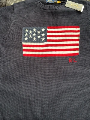 Mörkblå stickad Ralph Lauren tröja - Mörkblå stickad tröja från Polo Ralph Lauren med amerikansk flagga i rött, vitt och blått på bröstet samt RL-broderi. Tröjan har rund halsringning och ribbade muddar vid ärmslut och nederkant. Perfekt för dig som gillar klassisk och stilren design.