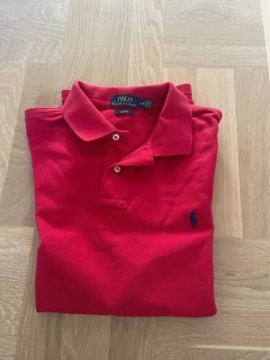 Röd pikétröja från Ralph Lauren - Klassisk röd pikétröja från Ralph Lauren med blå broderad logga på bröstet. Tröjan har krage, korta ärmar och knäppning med två knappar framtill. Perfekt för en stilren och sportig look.