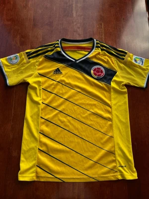 Gul Colombia Adidas fotbollströja - Gul Colombia fotbollströja från Adidas med svarta diagonala linjer och svarta detaljer på axlarna. Tröjan har rund hals med svart och vitt kantband, broderad landslagsemblem och Adidas-logga på bröstet. Tillverkad i lätt funktionsmaterial.