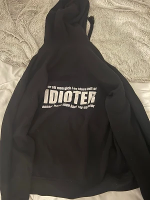 Svart hoodie Laröds Skola 2015 - Svart hoodie med tryck fram och bak. På bröstet står det 'Laröds Skola 2015' med ett emblem, och på ryggen finns ett stort tryck med texten 'IDIOTER'. Hoodien har huva med snören och en stor magficka. Perfekt för dig som vill ha en unik och personlig stil.