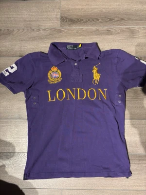Lila pikétröja från Ralph Lauren - Snygg lila pikétröja från Ralph Lauren med guldig broderad logga och texten LONDON på bröstet. Tröjan har korta ärmar, krage och knappar framtill samt stora siffror på ärm och rygg. Perfekt för en sportig och trendig look. Slim fit