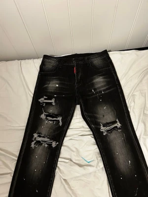 Svarta distressed jeans Dsquared2 - Säljer ett par svarta jeans från Dsquared2 med slitna detaljer, hål och vita färgstänk. Jeansen har smal passform, klassiska fem fickor och broderade blommor samt D2 på bakfickan. Perfekta för en edgy streetwear-look. Priset går och diskutera vid snabbköp 