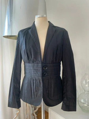 Svart kavaj med markerad midja - Stilren svart kavaj med två stora knappar och bred, veckad midjedetalj. Kavajen har långa ärmar, djup V-ringning och två framfickor. Materialet ser ut att vara glansigt, troligen polyester eller sidenmix. Snyggt skräddad passform som ger en cool siluett.