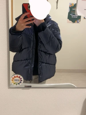 Mörkblå glansig pufferjacka - Snygg mörkblå pufferjacka med glansig finish och huva. Jackan har breda kanaler och dragkedja framtill. Perfekt för kalla dagar och ger en trendig look. Passar dig som vill ha en varm och stilren vinterjacka.