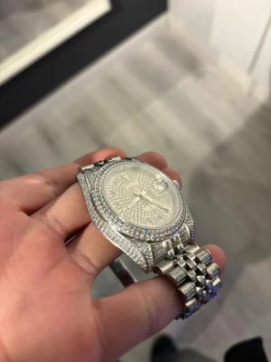 Silverfärgad klocka med stenar - Säljer en riktigt blingad klocka i silverfärg med massor av gnistrande stenar på både boett och armband. Klockan har en klassisk rund urtavla och länkarmband i metall. Perfekt för dig som vill sticka ut och gillar exklusiv stil. Gjord av Mossanite 